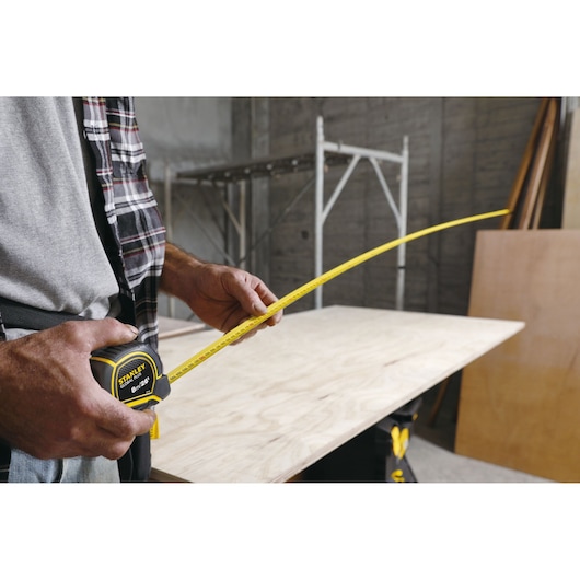 Photo of a person using a STANLEY 30-626 tape measure to measure a wooden board on a workbench.