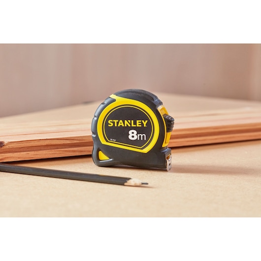 STANLEY 30-757 tape measure, 8m, black and yellow, on a workbench with pencil and wood planks, product photography.