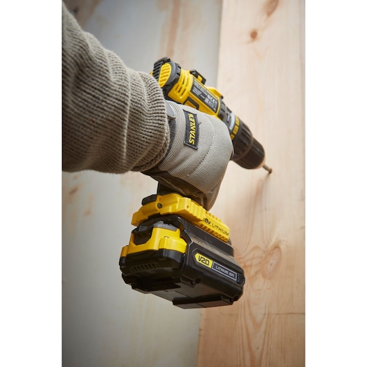 Stanley SBA100 cordless drill held by gloved hand, drilling into wood, side angle, yellow and black tool.