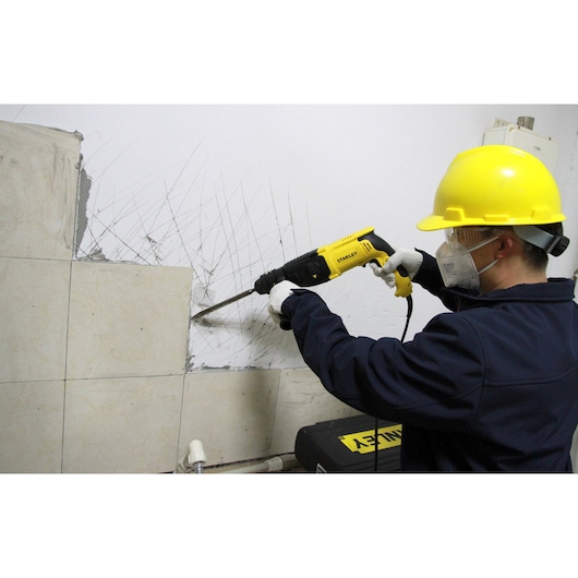 Stanley SHR264 rotary hammer drill in use removing wall tiles, side angle, yellow tool with black accents.
