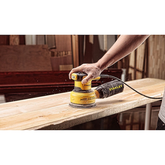 Photo of STANLEY SS30-B2C random orbital sander in use on wooden plank, handheld, dust bag attached.