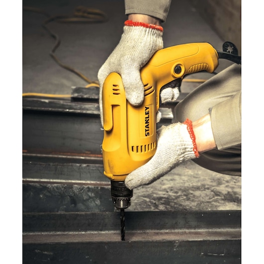 STANLEY STDR5510 yellow electric drill in use, drilling metal, product side angle, gloved hands visible.