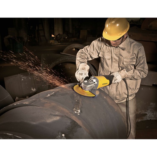 Photo of worker using STANLEY STGL2223 angle grinder on large metal pipe, producing visible sparks in industrial setting.