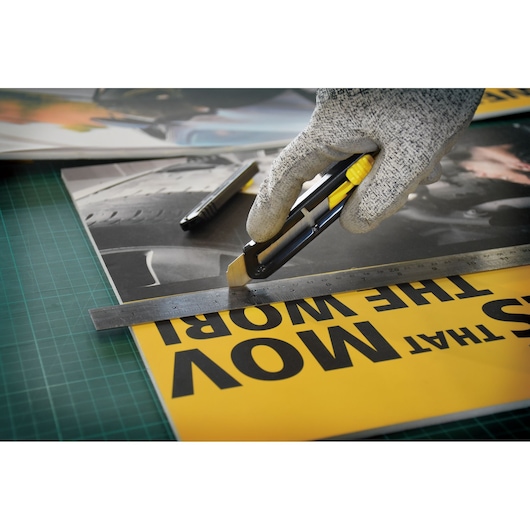 STANLEY STHT10323-840 utility knife cutting yellow poster along metal ruler, hand in gray glove holding knife.