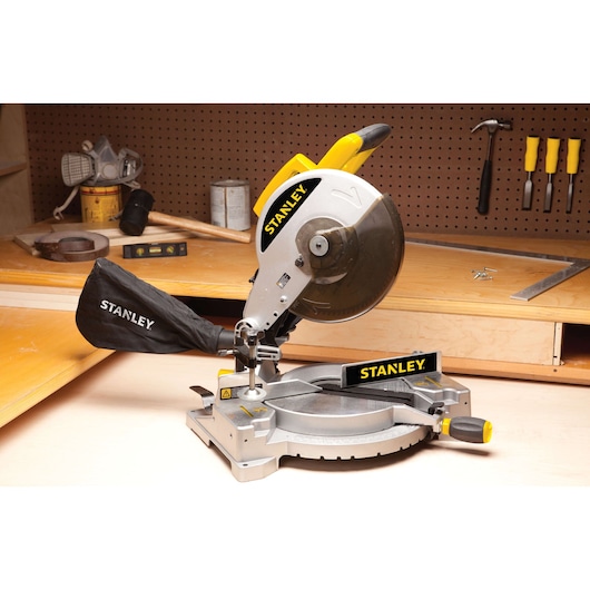 STANLEY STSM1525 miter saw with dust bag on workbench, silver and yellow, side angle, workshop background.